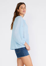 [Color: Light Blue] A side facing image of a brunette model wearing a light blue sheer bohemian blouse with long raglan sleeves, a round neckline, a button front, and a relaxed fit. 