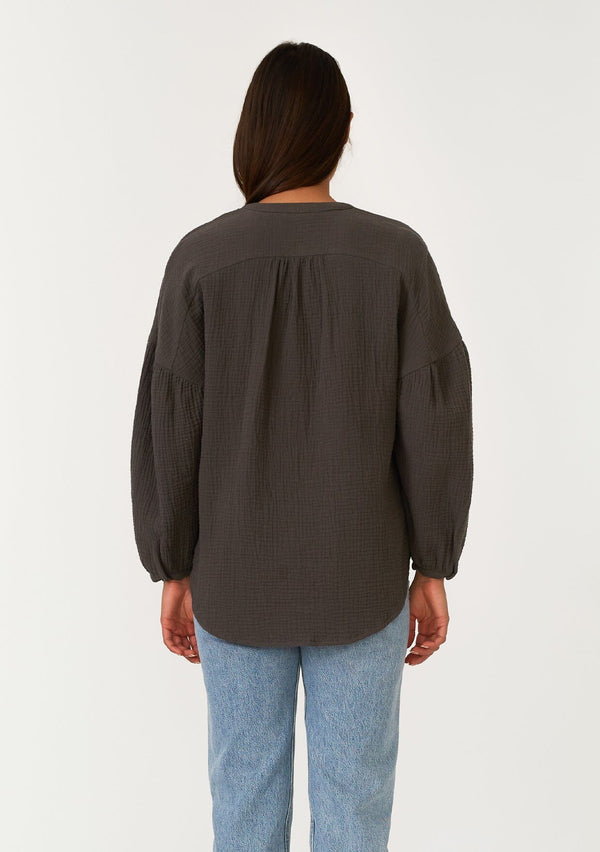 [Color: Lagoon] A back facing image of a brunette model wearing a classic bohemian blouse in an army green soft cotton gauze. With voluminous long sleeves, a dropped shoulder, a v neckline, a self covered button front, and a loose, relaxed fit. 