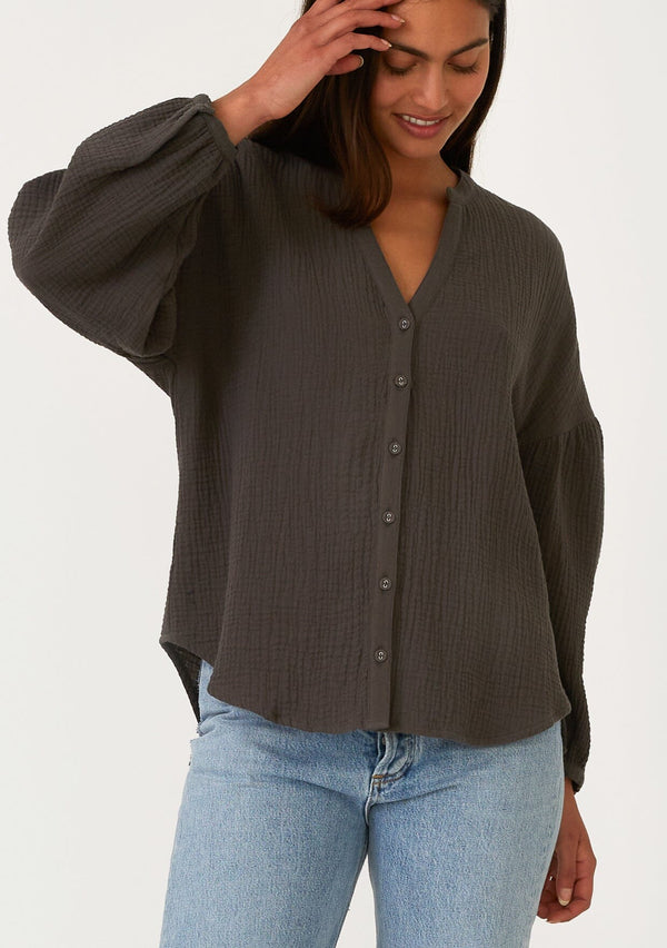 [Color: Lagoon] A front facing image of a brunette model wearing a classic bohemian blouse in an army green soft cotton gauze. With voluminous long sleeves, a dropped shoulder, a v neckline, a self covered button front, and a loose, relaxed fit. 