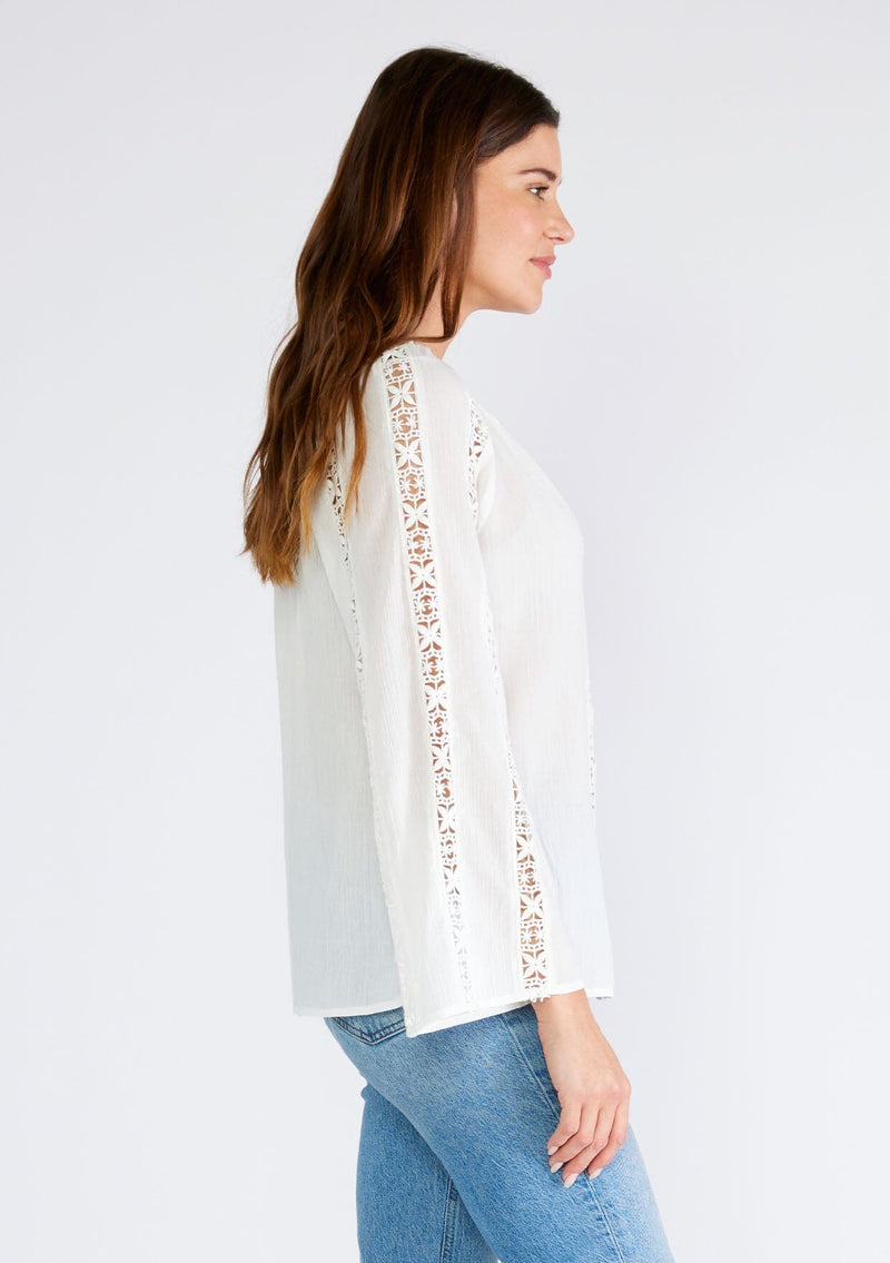 [Color: White] A side facing image of a brunette model wearing a classic bohemian white cotton blouse. With crochet trim, a split v neckline, and long bell sleeves. 