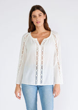 [Color: White] A half body front facing image of a brunette model wearing a classic bohemian white cotton blouse. With crochet trim, a split v neckline, and long bell sleeves. 