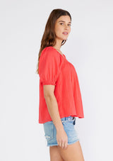 [Color: Hibiscus] A side facing image of a brunette model wearing a bright red cotton bohemian top with short puff sleeved, a ruffled elastic cuff, a square neckline, and lace trim. 