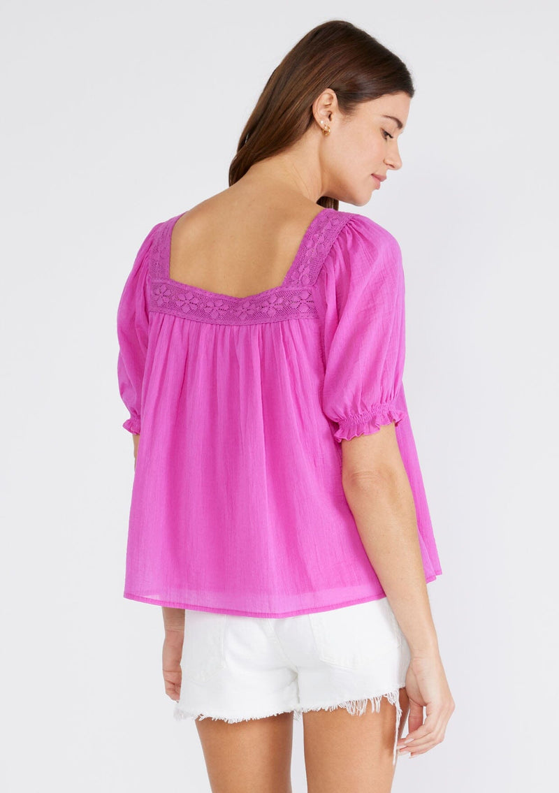 [Color: Orchid] A back facing image of a brunette model wearing a bright purple cotton bohemian top with short puff sleeved, a ruffled elastic cuff, a square neckline, and lace trim. 