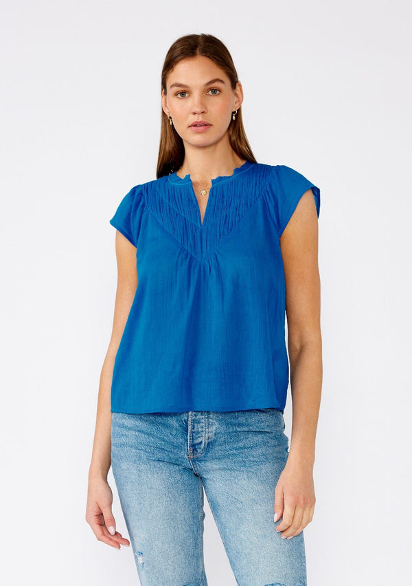 [Color: Cobalt] A front facing image of a brunette model wearing a bright blue bohemian top crafted from lightweight cotton gauze. With short cap sleeves, a split v neckline, gathered details at the yoke, and a raw edge hem. 
