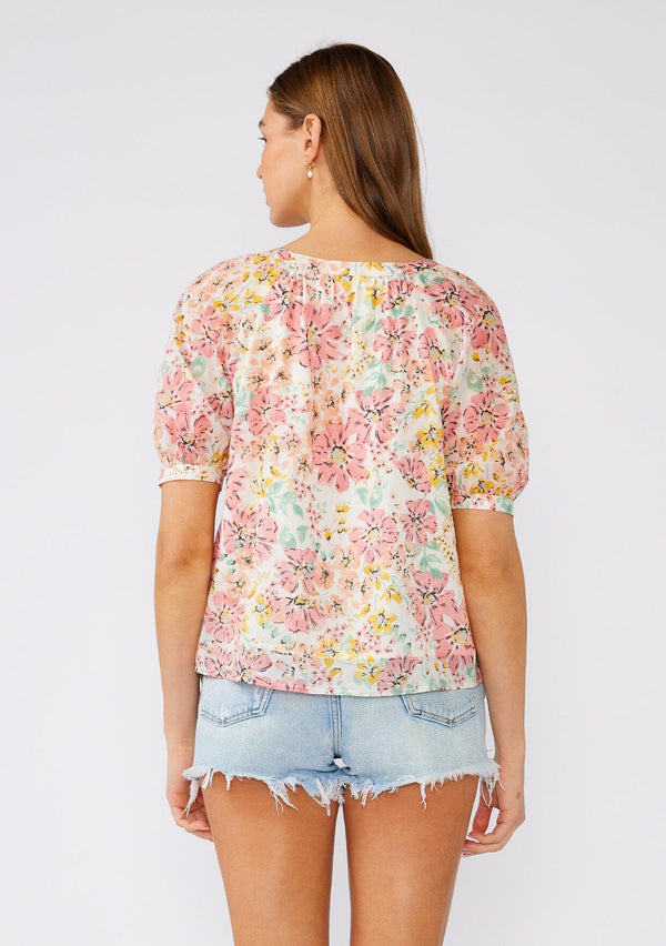[Color: Natural/Peach Blossom] A back facing image of a brunette model wearing a pink floral cotton summer blouse. With short puff sleeves, a scooped neckline, and a button front. 