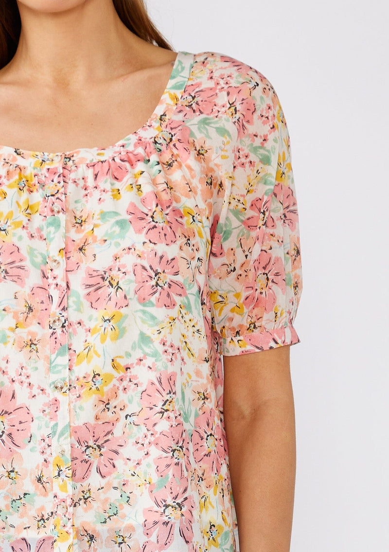 [Color: Natural/Peach Blossom] A close up front facing image of a brunette model wearing a pink floral cotton summer blouse. With short puff sleeves, a scooped neckline, and a button front. 