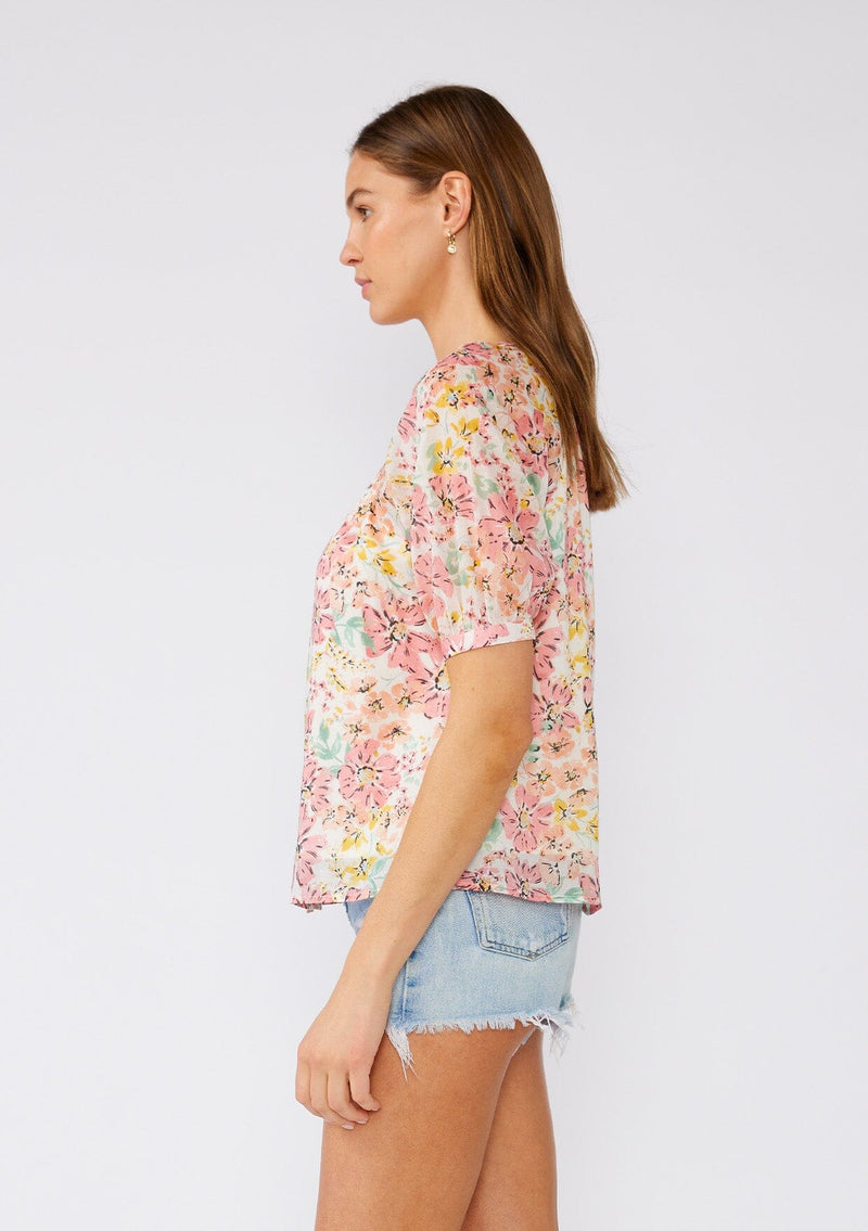 [Color: Natural/Peach Blossom] A side facing image of a brunette model wearing a pink floral cotton summer blouse. With short puff sleeves, a scooped neckline, and a button front. 
