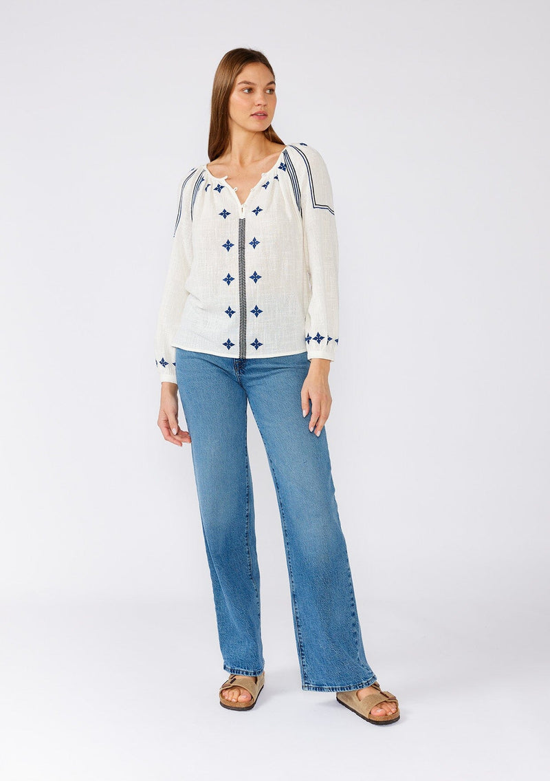 [Color: White/Navy] A full body front facing image of a brunette model wearing a white cotton bohemian blouse with navy blue embroidered details. With long sleeves, a round neckline, a button front, and a relaxed fit. 