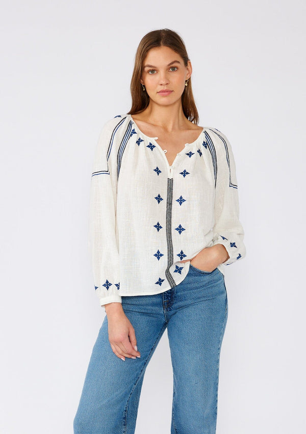 [Color: White/Navy] A front facing image of a brunette model wearing a white cotton bohemian blouse with navy blue embroidered details. With long sleeves, a round neckline, a button front, and a relaxed fit. 