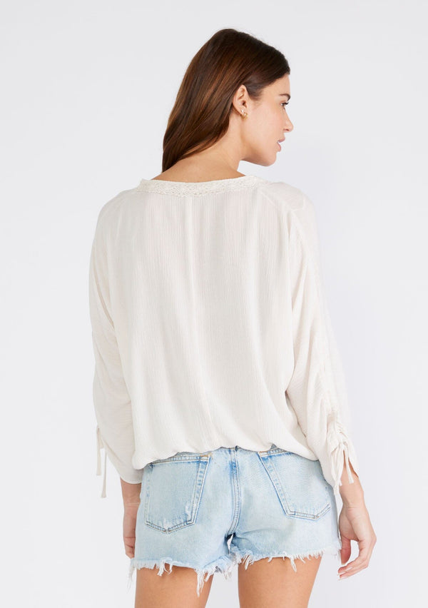 [Color: Almond] A back facing image of a brunette model wearing a light ivory bohemian blouse with three quarter length sleeves, a drawstring sleeve with tie accent, a v neckline, a self covered button front, and crochet lace trim. 