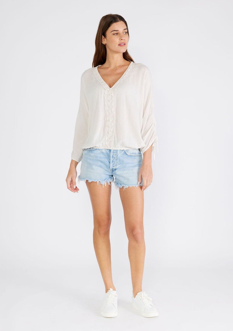 [Color: Almond] A full body front facing image of a brunette model wearing a light ivory bohemian blouse with three quarter length sleeves, a drawstring sleeve with tie accent, a v neckline, a self covered button front, and crochet lace trim. 