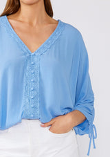 [Color: Periwinkle] A close up front facing image of a brunette model wearing a blue bohemian blouse with three quarter length sleeves, a drawstring sleeve with tie accent, a v neckline, a self covered button front, and crochet lace trim. 