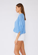 [Color: Periwinkle] A side facing image of a brunette model wearing a blue bohemian blouse with three quarter length sleeves, a drawstring sleeve with tie accent, a v neckline, a self covered button front, and crochet lace trim. 
