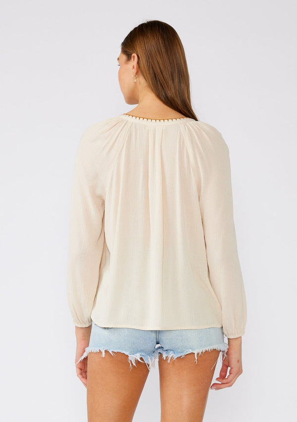 [Color: Natural/Pink] A back facing image of a brunette model wearing a cream colored bohemian blouse with floral embroidered details. With long raglan sleeves, a v neckline, a button front, and contrast stitch details along the hemline. 