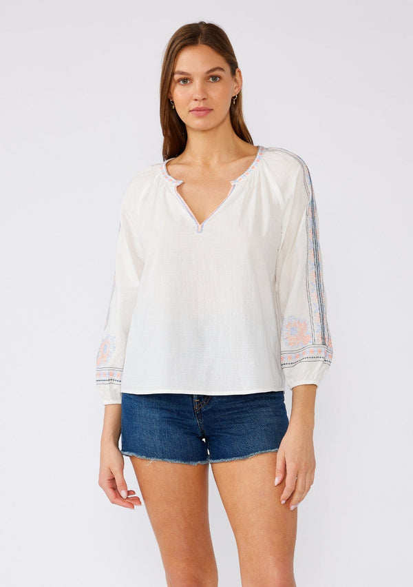 [Color: Off White/Blue] A front facing image of a brunette model wearing a white cotton bohemian blouse with colorful embroidered detail throughout. With three quarter length sleeves and a split v neckline. 