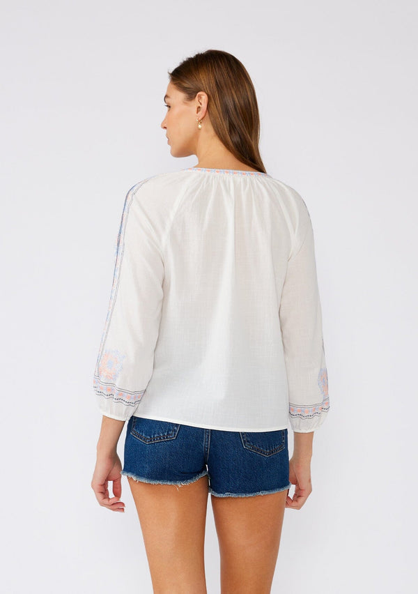 [Color: Off White/Blue] A back facing image of a brunette model wearing a white cotton bohemian blouse with colorful embroidered detail throughout. With three quarter length sleeves and a split v neckline. 