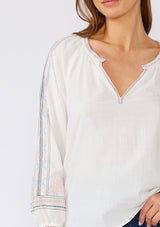 [Color: Off White/Blue] A close up front facing image of a brunette model wearing a white cotton bohemian blouse with colorful embroidered detail throughout. With three quarter length sleeves and a split v neckline. 