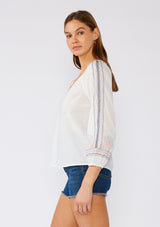 [Color: Off White/Blue] A side facing image of a brunette model wearing a white cotton bohemian blouse with colorful embroidered detail throughout. With three quarter length sleeves and a split v neckline. 