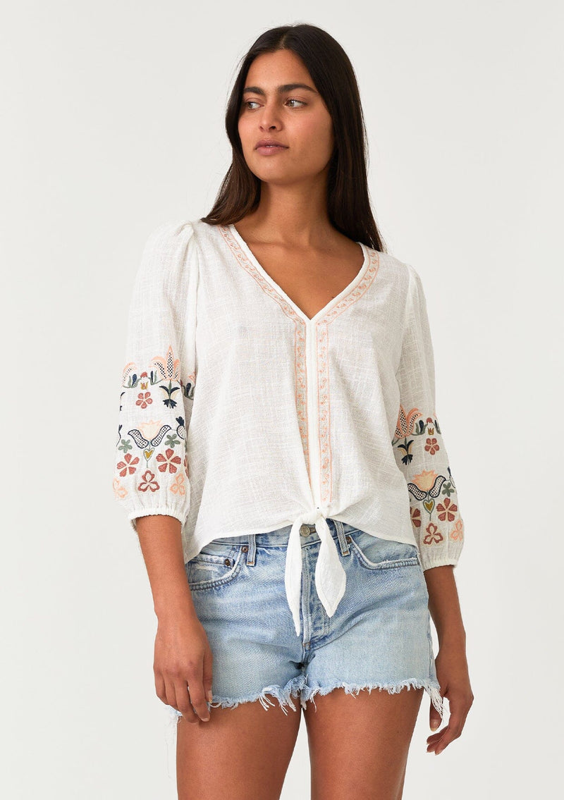 [Color: Natural/Coral] A front facing image of a brunette model wearing an off white bohemian blouse with colorful floral embroidered details throughout. Featuring three quarter length sleeves, a v neckline, and a tie waist detail. 