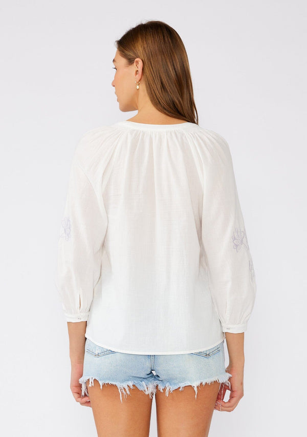 [Color: Off White/Dusty Lavender] A back facing image of a brunette model wearing a lightweight white cotton bohemian blouse with light purple floral embroidered detail. With three quarter length sleeves, a round neckline with smocked details, and a self covered button front. 