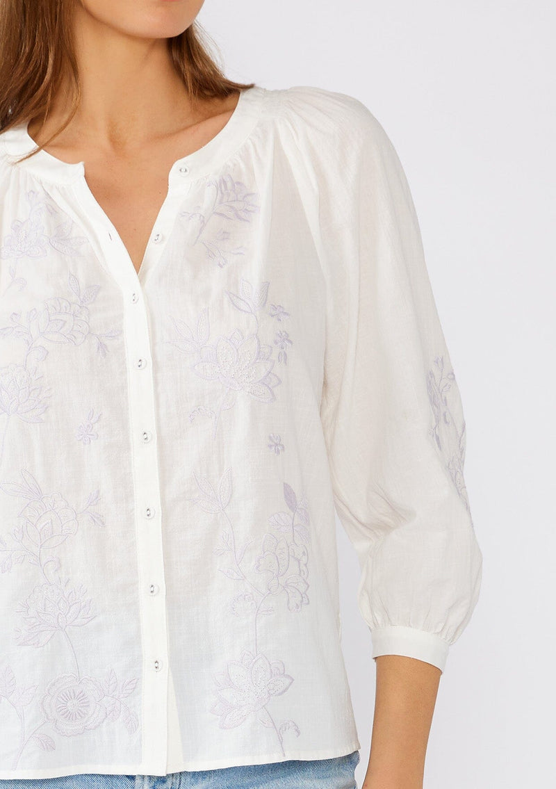 [Color: Off White/Dusty Lavender] A close up front facing image of a brunette model wearing a lightweight white cotton bohemian blouse with light purple floral embroidered detail. With three quarter length sleeves, a round neckline with smocked details, and a self covered button front. 