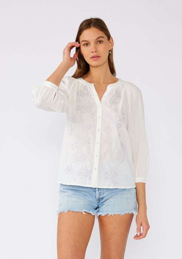 [Color: Off White/Dusty Lavender] A front facing image of a brunette model wearing a lightweight white cotton bohemian blouse with light purple floral embroidered detail. With three quarter length sleeves, a round neckline with smocked details, and a self covered button front. 
