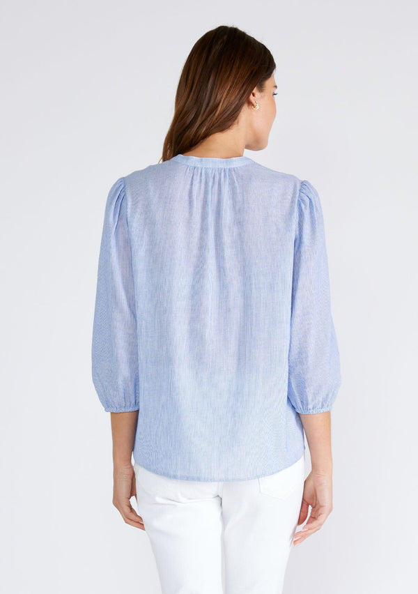 [Color: Chambray/Ivory] A back facing image of a brunette model wearing a classic cotton blouse in a blue and white pinstripe. With three quarter length sleeves, a split v neckline, a button front, pleated pintuck details, and contrasting blanket stitch details. The flowy fit is both comfortable and chic for spring. 