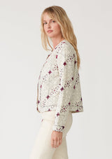 [Color: Natural/Fuchsia] A side facing image of a blonde model wearing a lightweight bohemian spring jacket in ivory with pink embroidered detail and contrast black thread detail. With long sleeves, an open front, and a cropped fit. 
