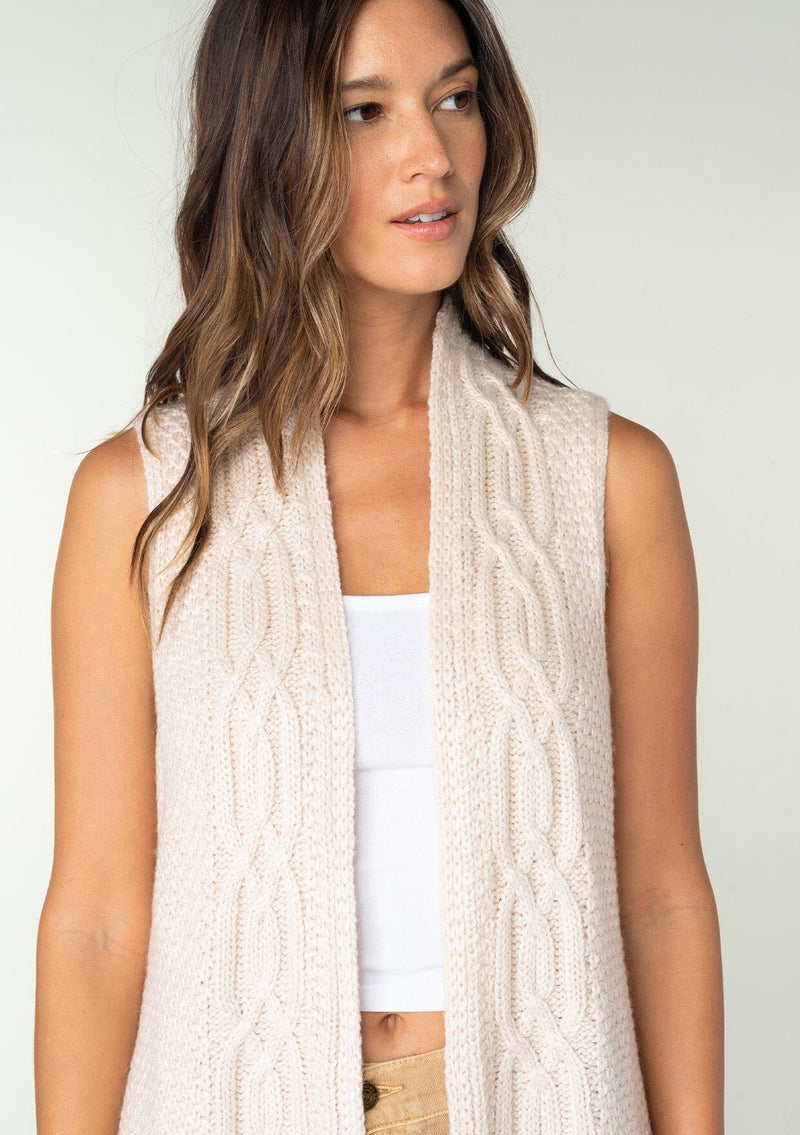 [Color: Oatmeal] A model wearing a cream colored chunky cable knit sweater vest. With a long mid length, side slits, and an open front.