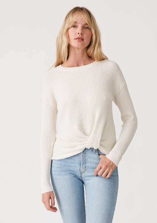 [Color: Natural] A front facing image of a blonde model wearing an ivory waffle knit pullover sweater. With long sleeves, a wide crew neckline, and a twist front detail at the waist. 