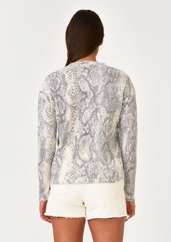 [Color: Light Grey/Ivory] A back facing image of a brunette model wearing a soft waffle knit pullover sweater in a blue grey and ivory snakeskin print. With long sleeves, a crew neckline, and a twist front detail at the waist. 