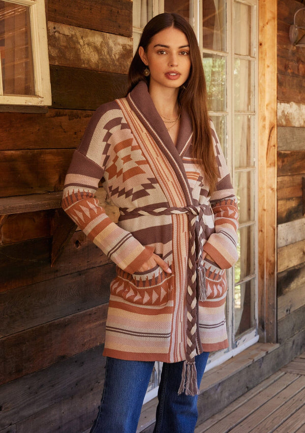 [Color: Taupe/Blush] A front facing image of a brunette model standing outside wearing a cozy fall bohemian cardigan in a pink and brown southwestern style pattern. With long sleeves, a shawl collar, side pockets, and a braided waist belt.