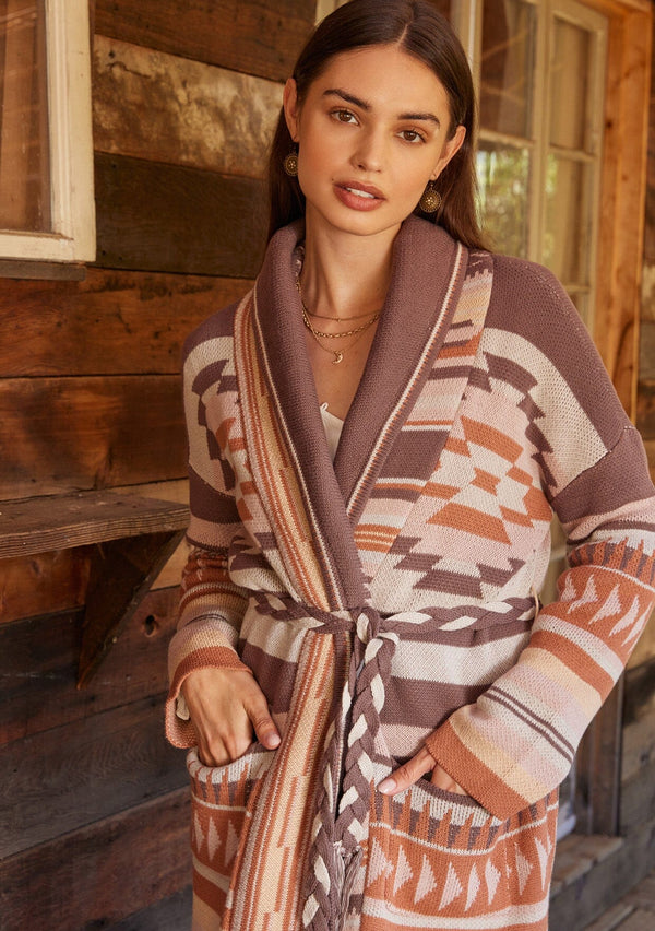 [Color: Taupe/Blush] A close up front facing image of a brunette model standing outside wearing a cozy fall bohemian cardigan in a pink and brown southwestern style pattern. With long sleeves, a shawl collar, side pockets, and a braided waist belt.