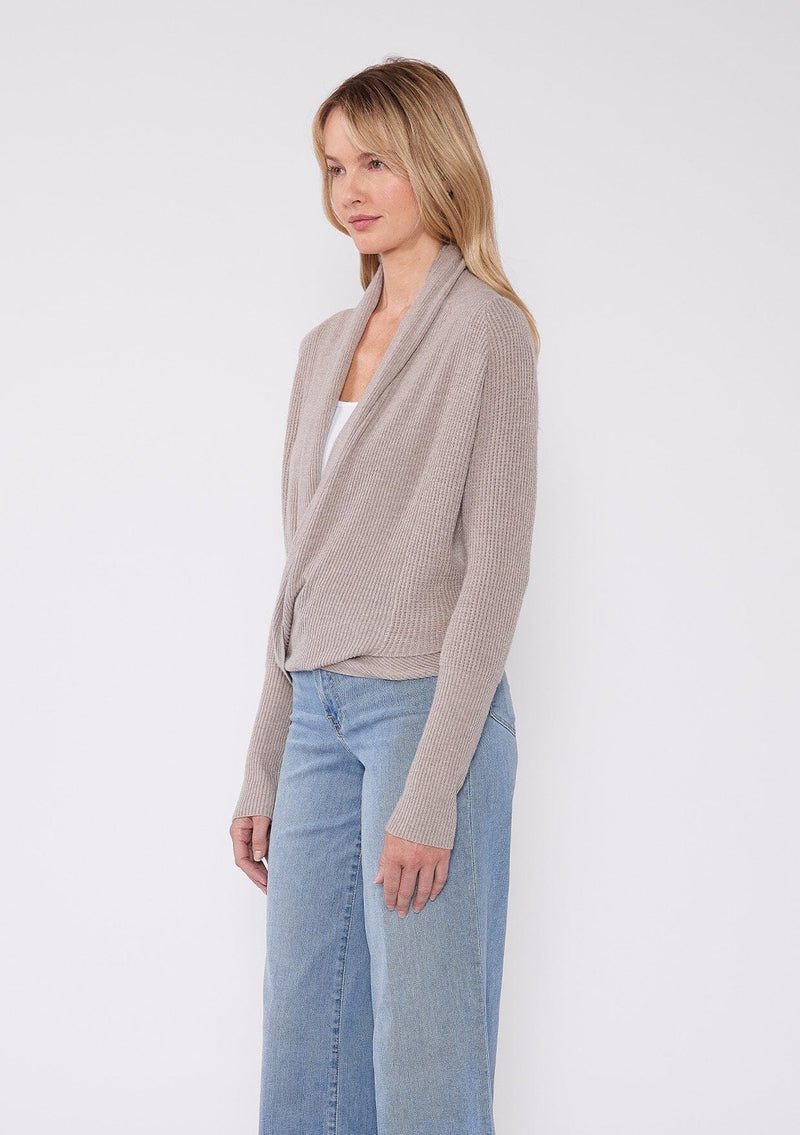 [Color: Heather Cement] A photo of a Lovestitch model wearing a chic beige waffle knit wrap front sweater with long sleeves, a v neckline, and a button closure at the back. The long ties can be styled in multiple ways.