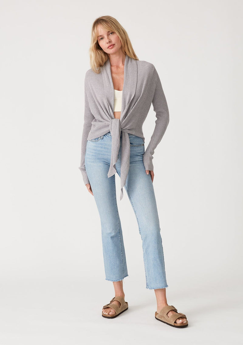 [Color: Heather Grey] A full body front facing image of a blonde model wearing a heather grey waffle knit wrap sweater with long sleeves, a v neckline, and a button closure at the back. The long ties can be styled in multiple ways. 