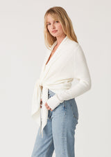 [Color: Ivory] A side facing image of a blonde model wearing an ivory waffle knit wrap sweater with long sleeves, a v neckline, and a button closure at the back. The long ties can be styled in multiple ways. 