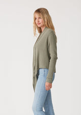 [Color: Olive] A side facing image of a blonde model wearing an olive green waffle knit wrap sweater with long sleeves, a v neckline, and a button closure at the back. The long ties can be styled in multiple ways. 