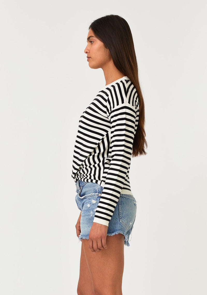 [Color: Cream/Black] A side facing image of a brunette model wearing a classic black and cream striped knit pullover sweater. With long sleeves, a drop shoulder, a crew neckline, and a twist front detail at the waist. 