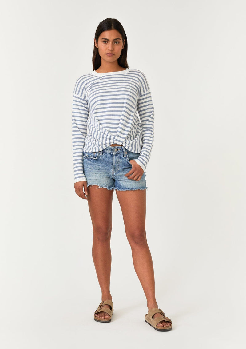 [Color: Ivory/Dusty Blue] A full body front facing image of a brunette model wearing a classic ivory and light blue striped knit pullover sweater. With long sleeves, a drop shoulder, a crew neckline, and a twist front detail at the waist. 