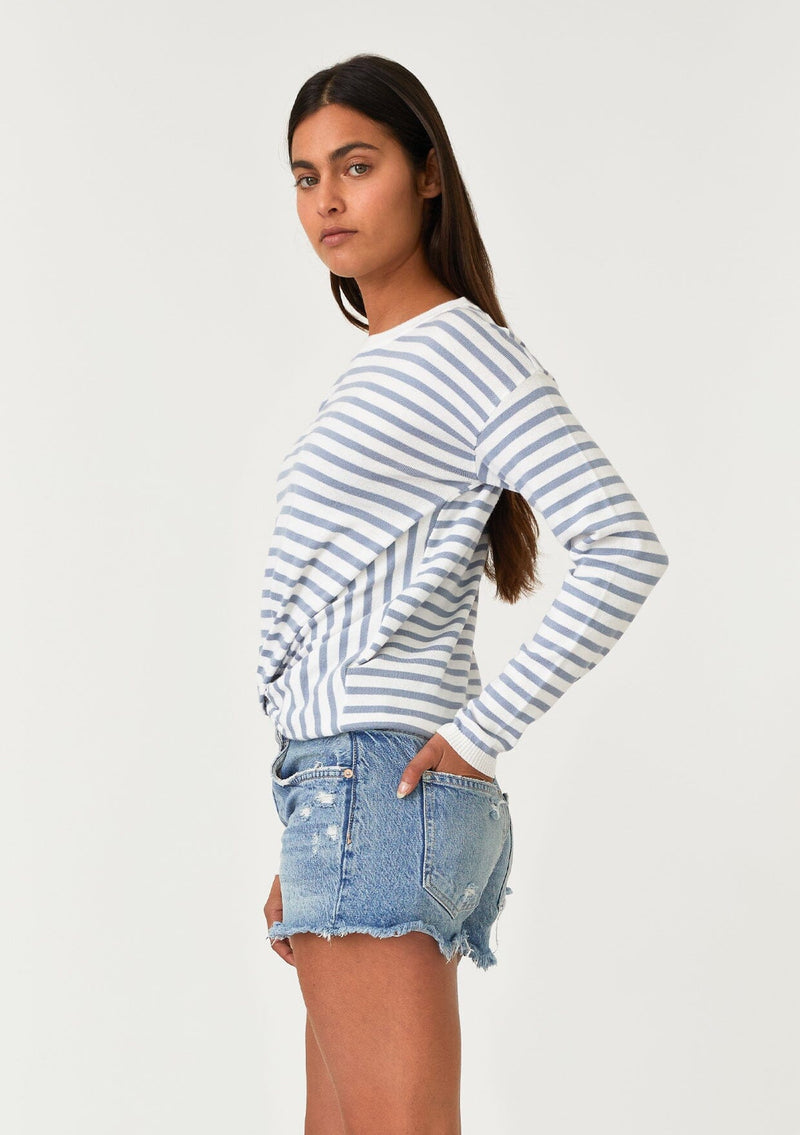 [Color: Ivory/Dusty Blue] A side facing image of a brunette model wearing a classic ivory and light blue striped knit pullover sweater. With long sleeves, a drop shoulder, a crew neckline, and a twist front detail at the waist. 