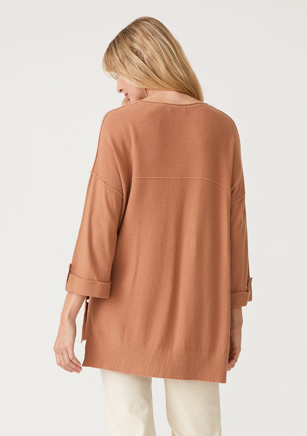 [Color: Clay] A back facing image of a blonde model wearing a clay brown lightweight sweater tunic top. With three quarter length sleeves, a button roll tab, a v neckline, exposed seam details, a long tunic length, and side vents. 