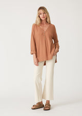 [Color: Clay] A full body front facing image of a blonde model wearing a clay brown lightweight sweater tunic top. With three quarter length sleeves, a button roll tab, a v neckline, exposed seam details, a long tunic length, and side vents. 