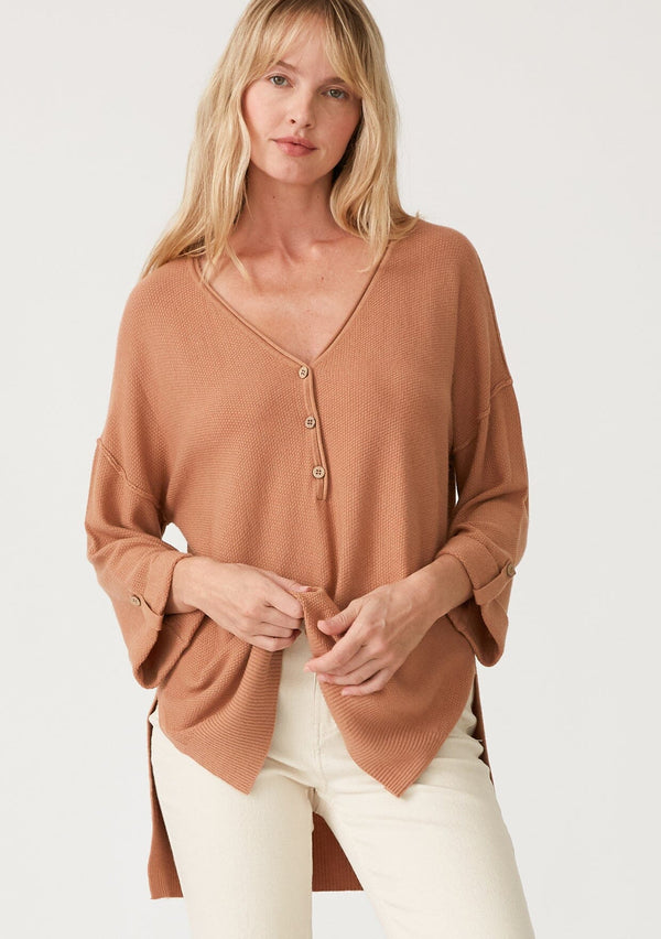 [Color: Clay] A front facing image of a blonde model wearing a clay brown lightweight sweater tunic top. With three quarter length sleeves, a button roll tab, a v neckline, exposed seam details, a long tunic length, and side vents. 