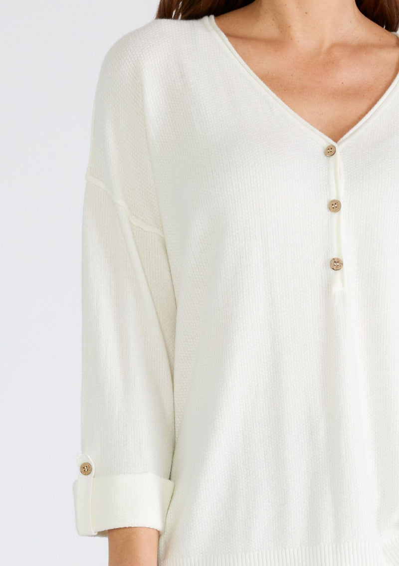 [Color: Cream] A close up front facing image of a brunette model wearing an ivory lightweight sweater tunic top. With three quarter length sleeves, a button roll tab, a v neckline, exposed seam details, a long tunic length, and side vents.