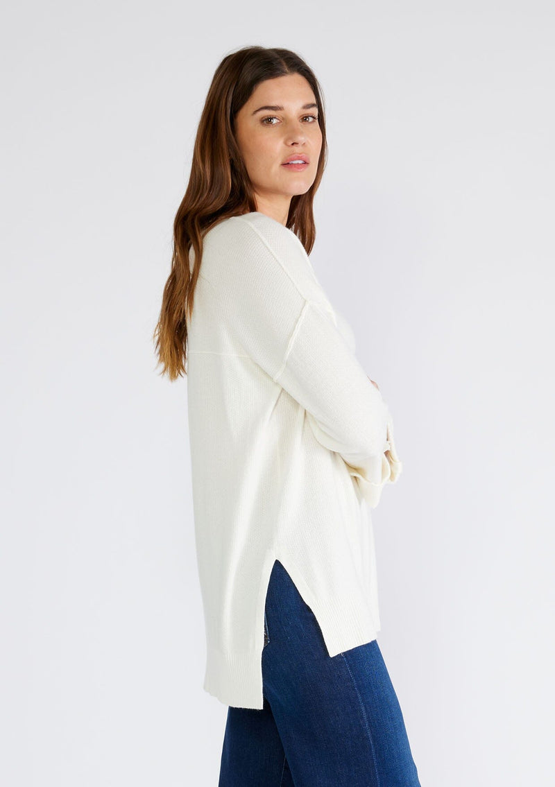 [Color: Cream] A side facing image of a brunette model wearing an ivory lightweight sweater tunic top. With three quarter length sleeves, a button roll tab, a v neckline, exposed seam details, a long tunic length, and side vents.