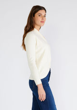 [Color: Cream] A side facing image of a brunette model wearing a cream colored knit sweater top. With a slim fit, long sleeves, a deep v neckline, and a twist front detail. 
