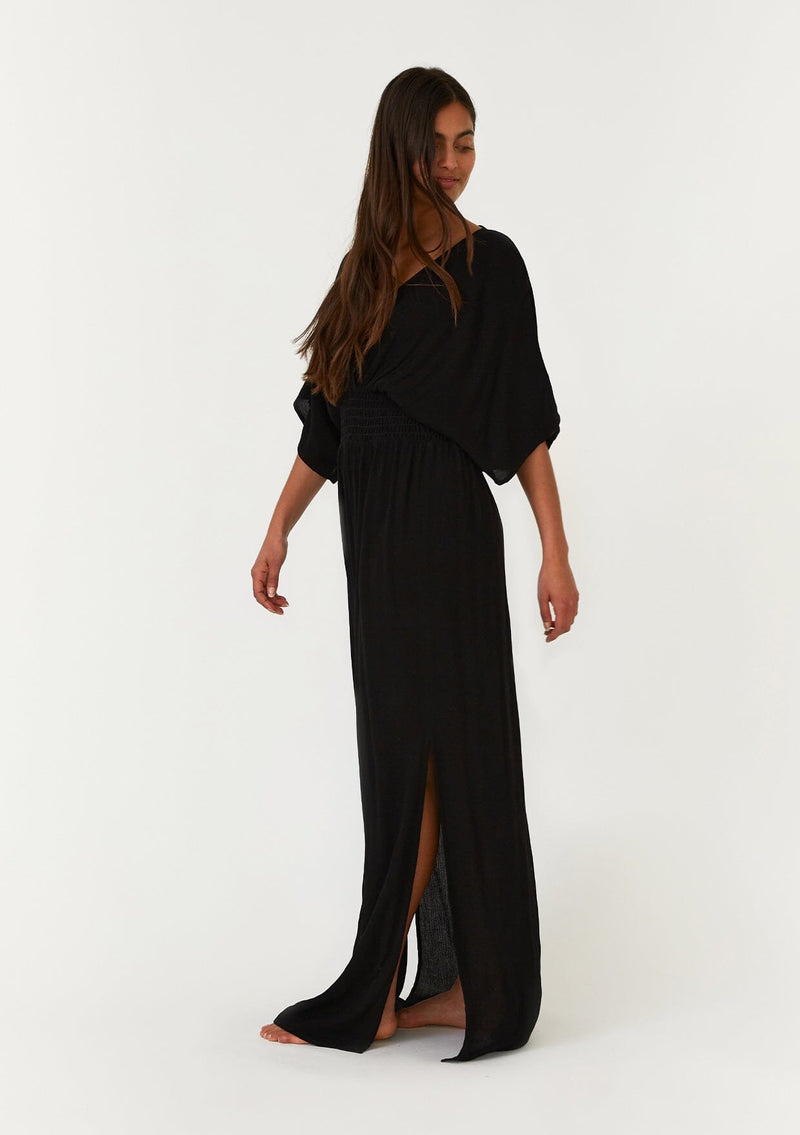 [Color: Black] A side facing image of a brunette model wearing a resort ready black maxi dress. With half length kimono sleeves, a plunging v neckline, a smocked elastic empire waist, side slits, and an open back with tie closure.