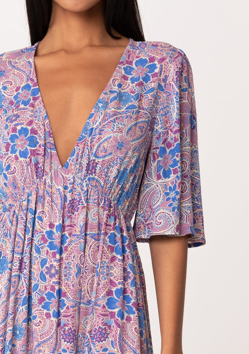 [Color: Dusty Rose/Blue] A close up front facing image of a brunette model wearing a flowy summer mini dress with half length sleeves, a v neckline, and an empire waist. Designed in a pink and blue floral print with gold clip dot details throughout. 