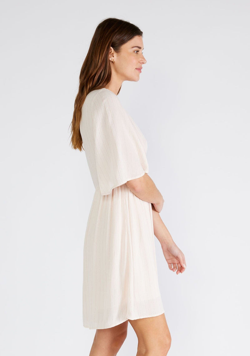 [Color: Light Peach] A side facing image of a brunette model wearing a classic flowy mini dress in light peach with a gold metallic thread detail. With short half length sleeves, a v neckline, and an empire waist.