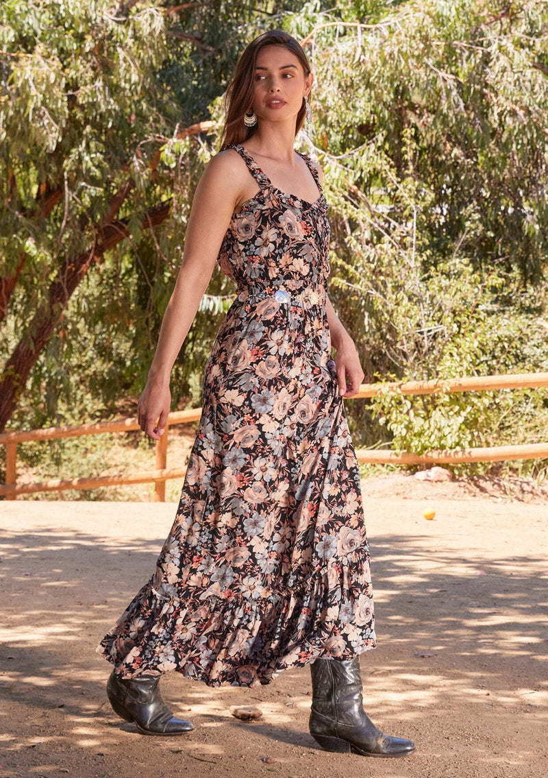 [Color: Black/Dusty Blue] A full body side facing image of a brunette model standing outside wearing a bohemian sleeveless maxi dress in a black and dusty blue floral print. With ruffled tank top straps, a tiered long skirt, an elastic waist, a square neckline, and a lace up tie front top.